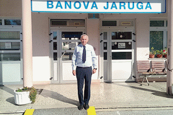 Željko Basić: Željko Basić u svijetloplavoj košulji s tamnoplavom kravatom stoji ispred kolodvorske zgrade, ispod natpisa Banova Jaruga, na sunčanom danu.