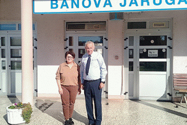 Mirjana Jozić u smeđoj odjeći i Željko Basić u svijetloplavoj košulji s kravatom stoje jedno uz drugo pred ulazom u kolodvor, iznad njih plavo-bijeli natpis Banova Jaruga.